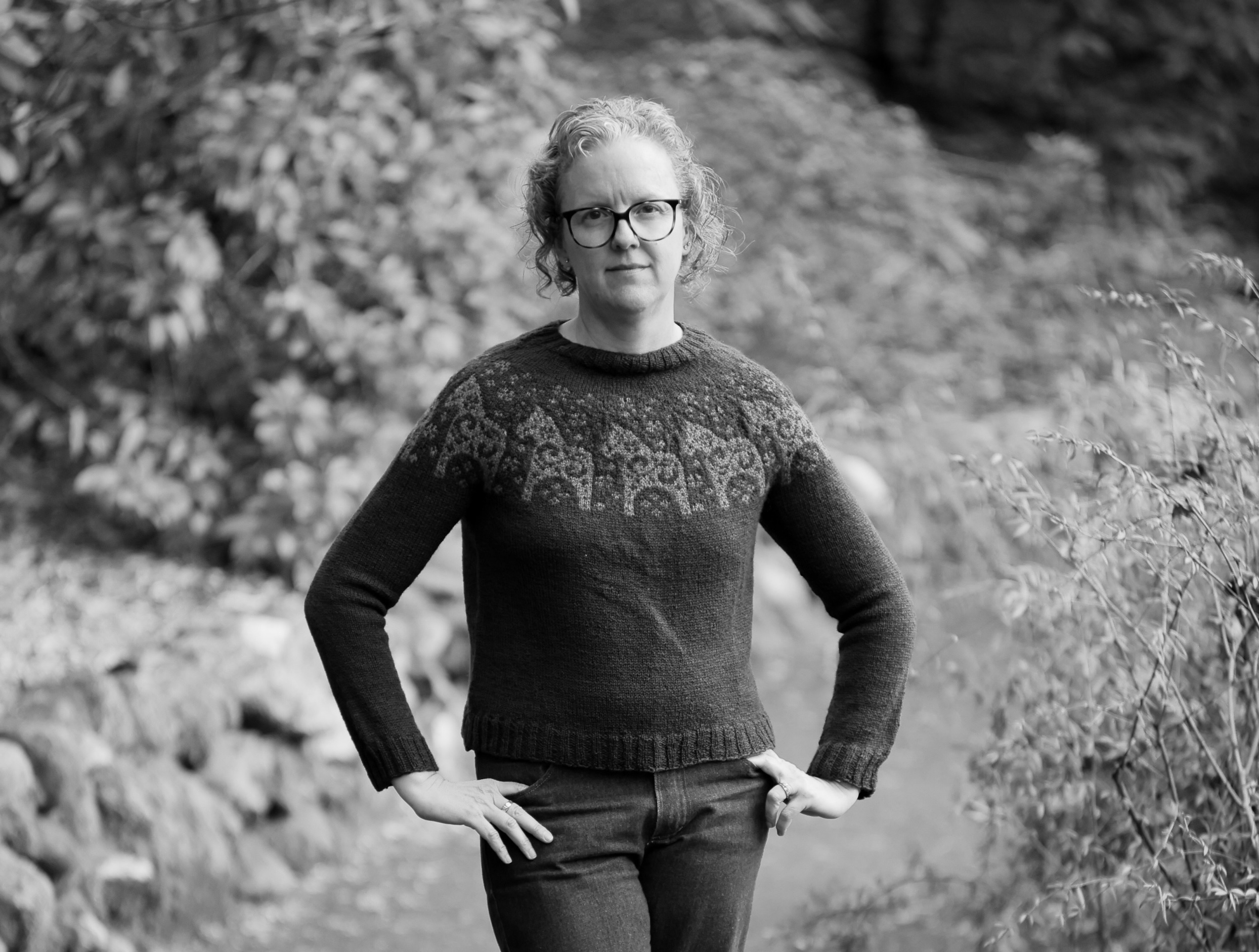 A woman stands on a pathway with fuzzy out of focus trees and bushes around and behind her. She's got her hands on her hips and stares directly at the camera. She's got chin length curly hair and is wearing dark framed glasses. She wears a sweater with a pattern of horses across the yoke and jeans. The photo is in black and white.