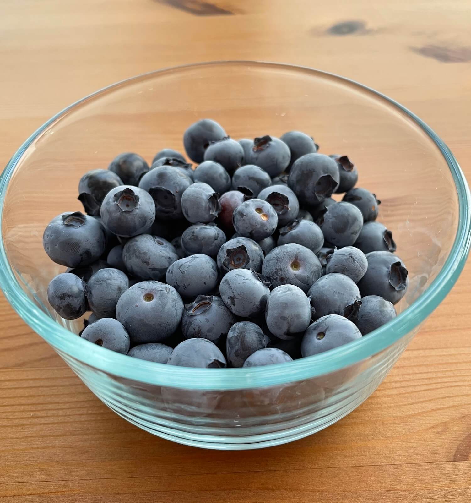 a clear glass bowl sitting on a wood table almost full of blueberries