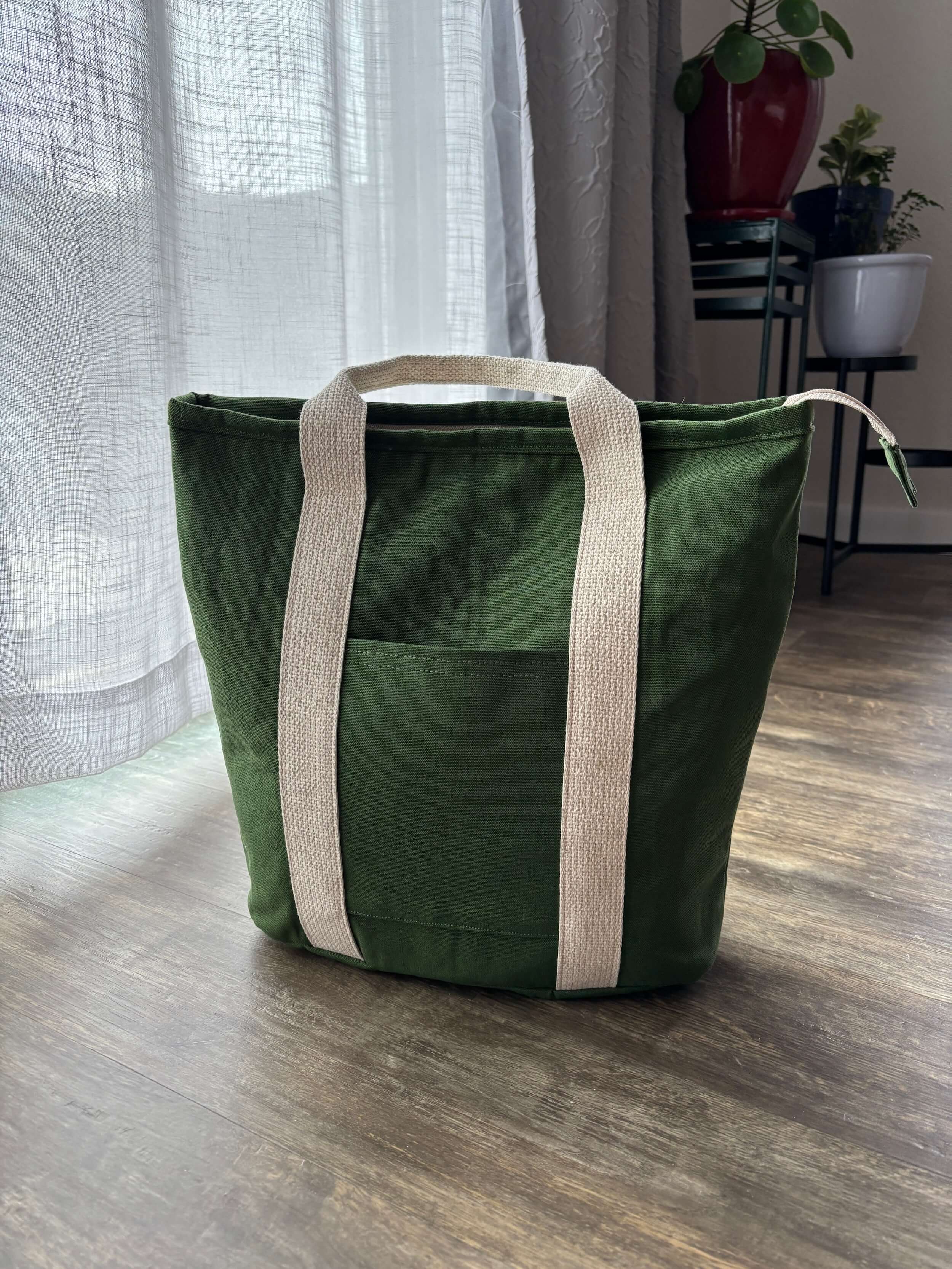 A green back sits on a brown floor in front of sheer white curtains. In the background are three plants that are slightly out of focus. The bag has wide cream colored straps and a front pocket. The zipper is longer than the top of the bag and hangs over the side.