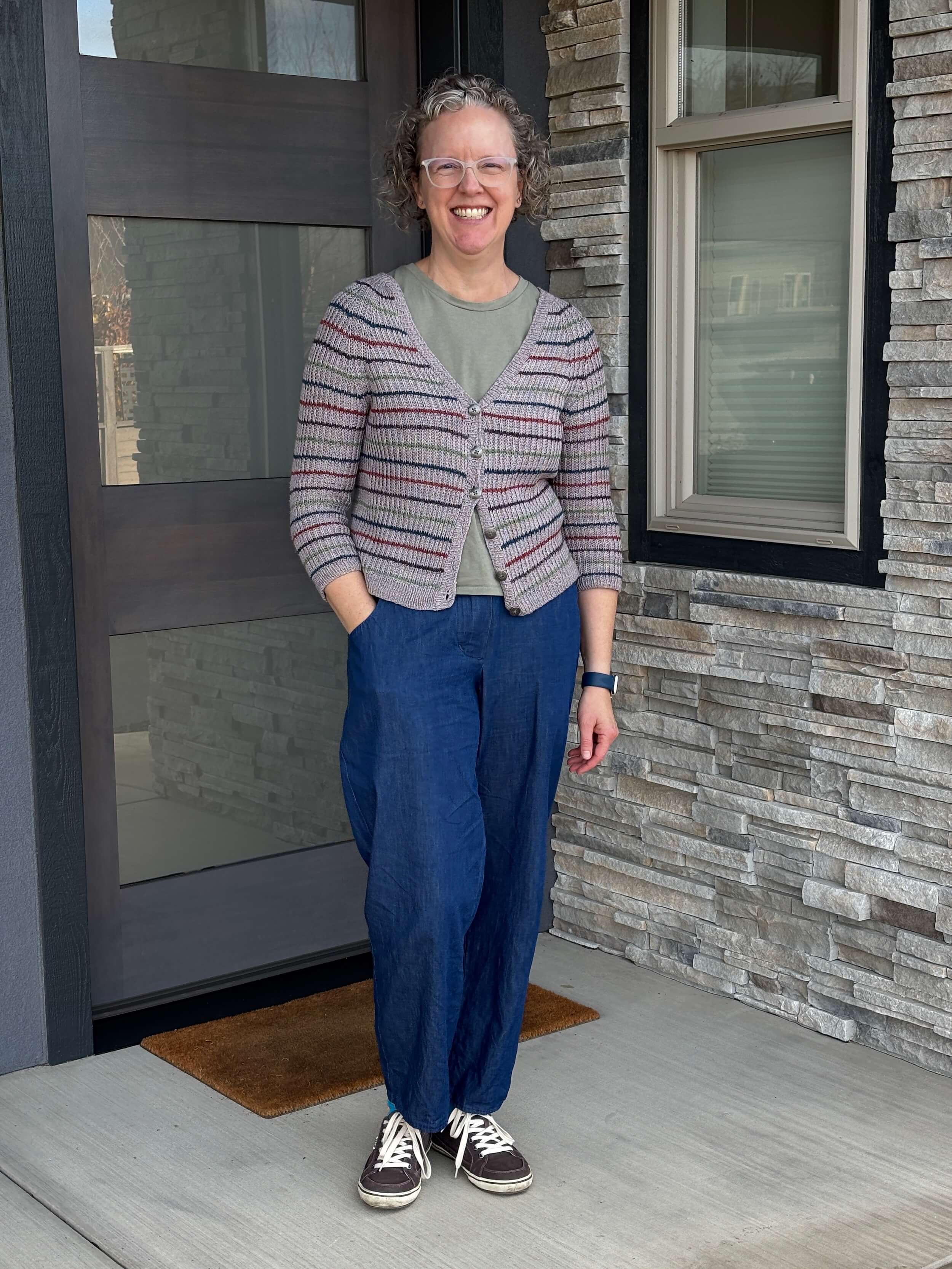 A woman stands in front of a doorway with her right hand in the pocket of her trousers, she's smiling, almost laughing, and she's wearing white plastic frame glasses. Her hair is curly gray blond, she's wearing a striped gray cardigan, a green t-shirt showing underneath, and blue wide leg denim trousers with blue and orange socks and gray brown tennis shoes.