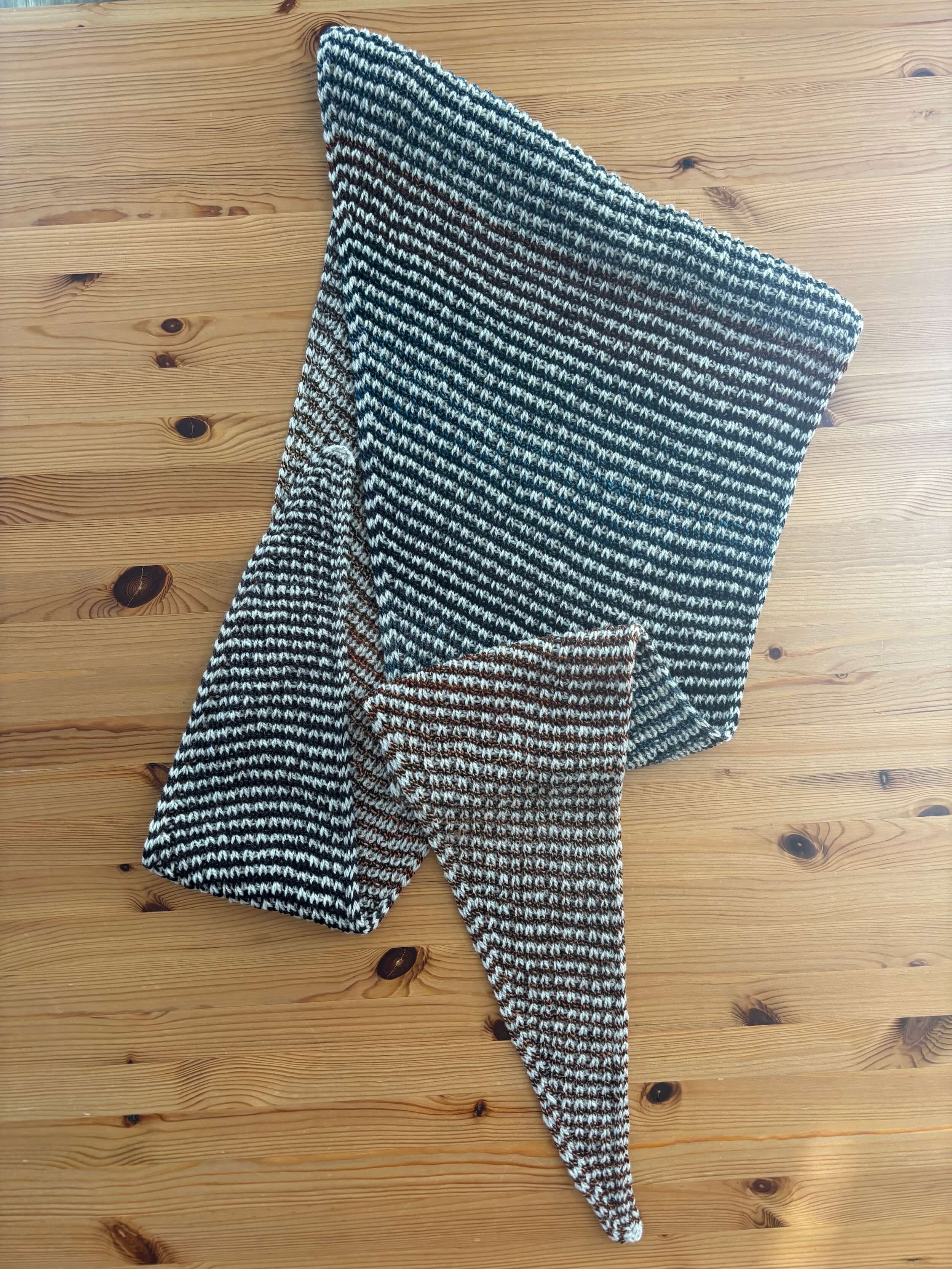 On a wood table where the wood grain shows, a scarf is laid out and doubled up. The scarf is striped with a light, natural color and a yarn that changes from dark to orange to blue.