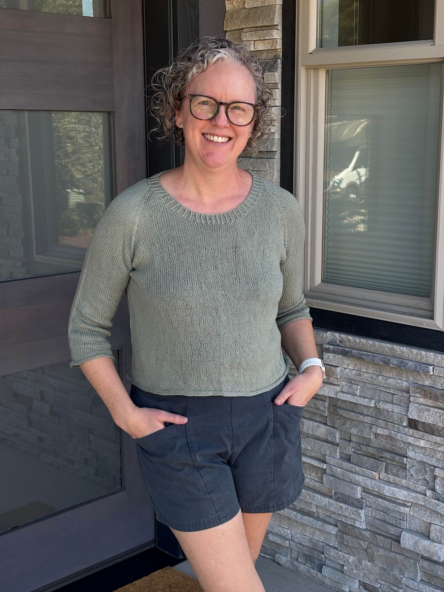 A woman stands in front of a doorway wearing a light green sweater that has three quarter length sleeves and a slight boxy fit. Her hands on in the pockets of her navy blue shorts and her legs are crossed over each other at the top of the thighs that you can see. She's smiling and has curly dirty blond hair and is wearing dark brown glasses.