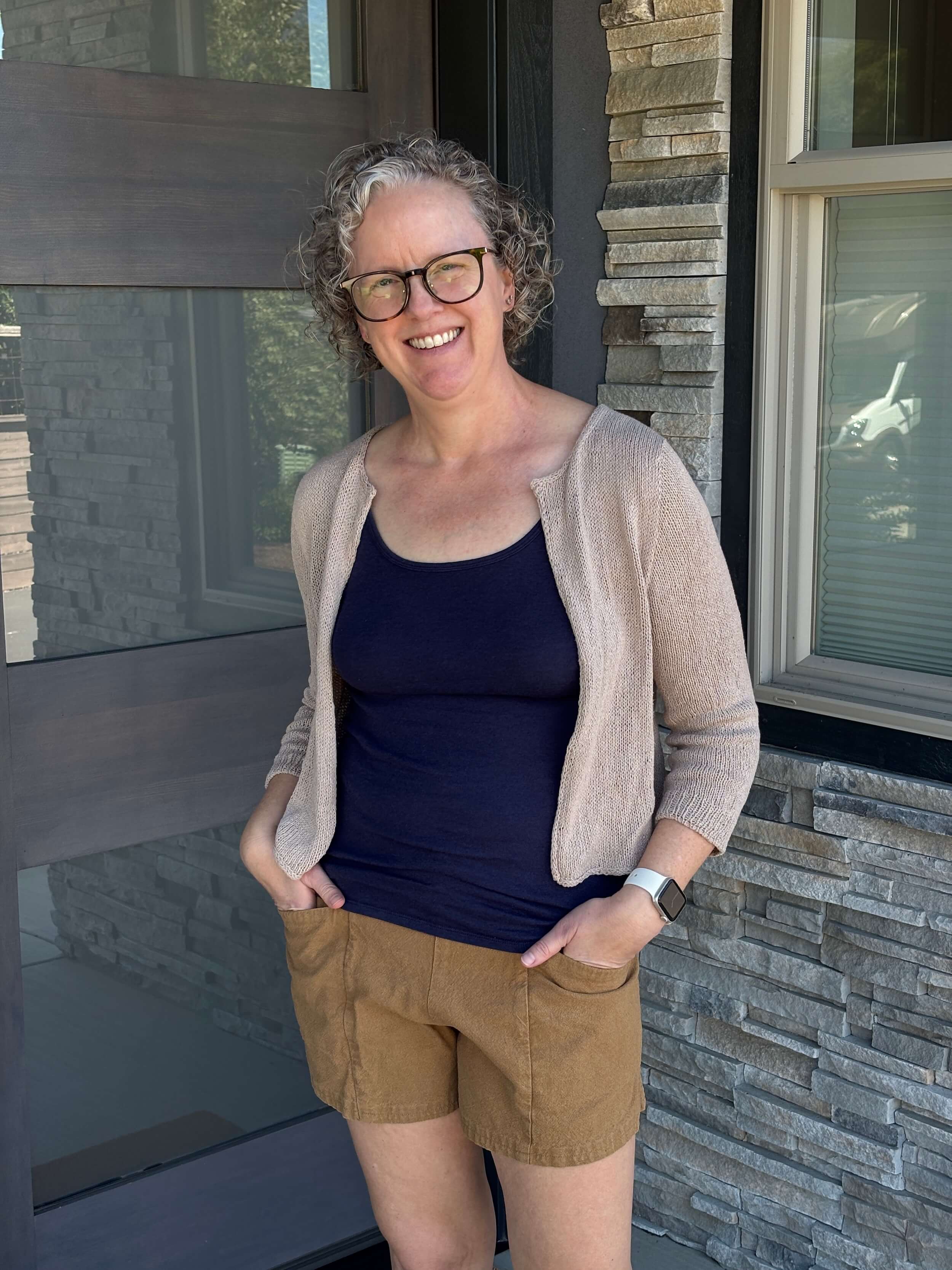 A woman stands in front of a doorway wearing a light brown cardigan sweater that is open at the front and doesn't have any buttons. Under the seater is a navy tank top and she's got her hands in the pockets of her pecan colored shorts. The sweater has three quarter length sleeves and hits at her hip bone. She's smiling, with her body angled to her right with her head slightly tilted to the right as well. She's got curly dirty blond hair and dark brown glasses on.