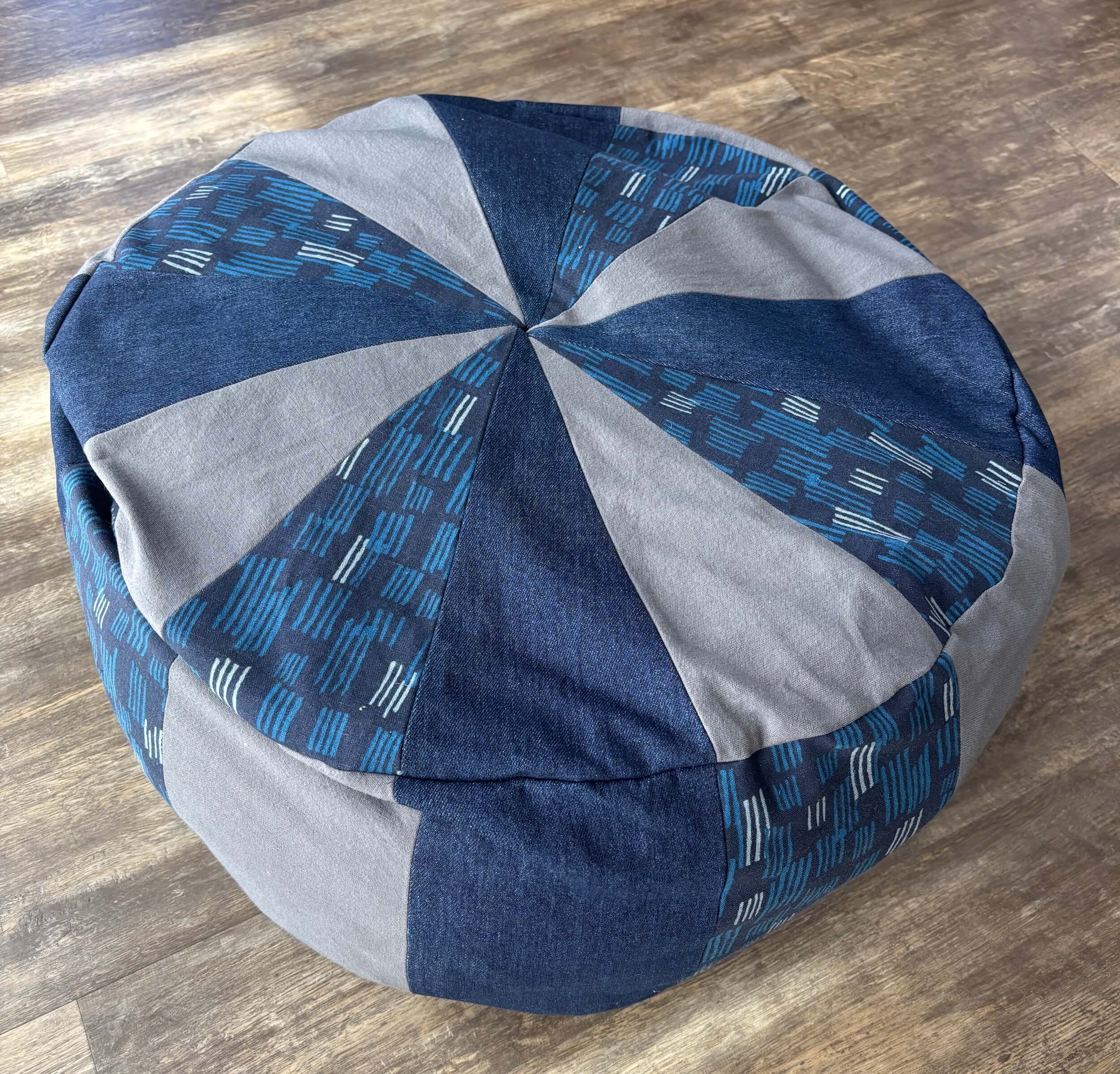A round cushion sits on a brown floor. The cushion has trangular pieces of fabric in blue denim, gray, and a patterned blue and white and dark blue canvas on the top, looking like a pie shape. On the sides are rectangular pieces in the same fabric.