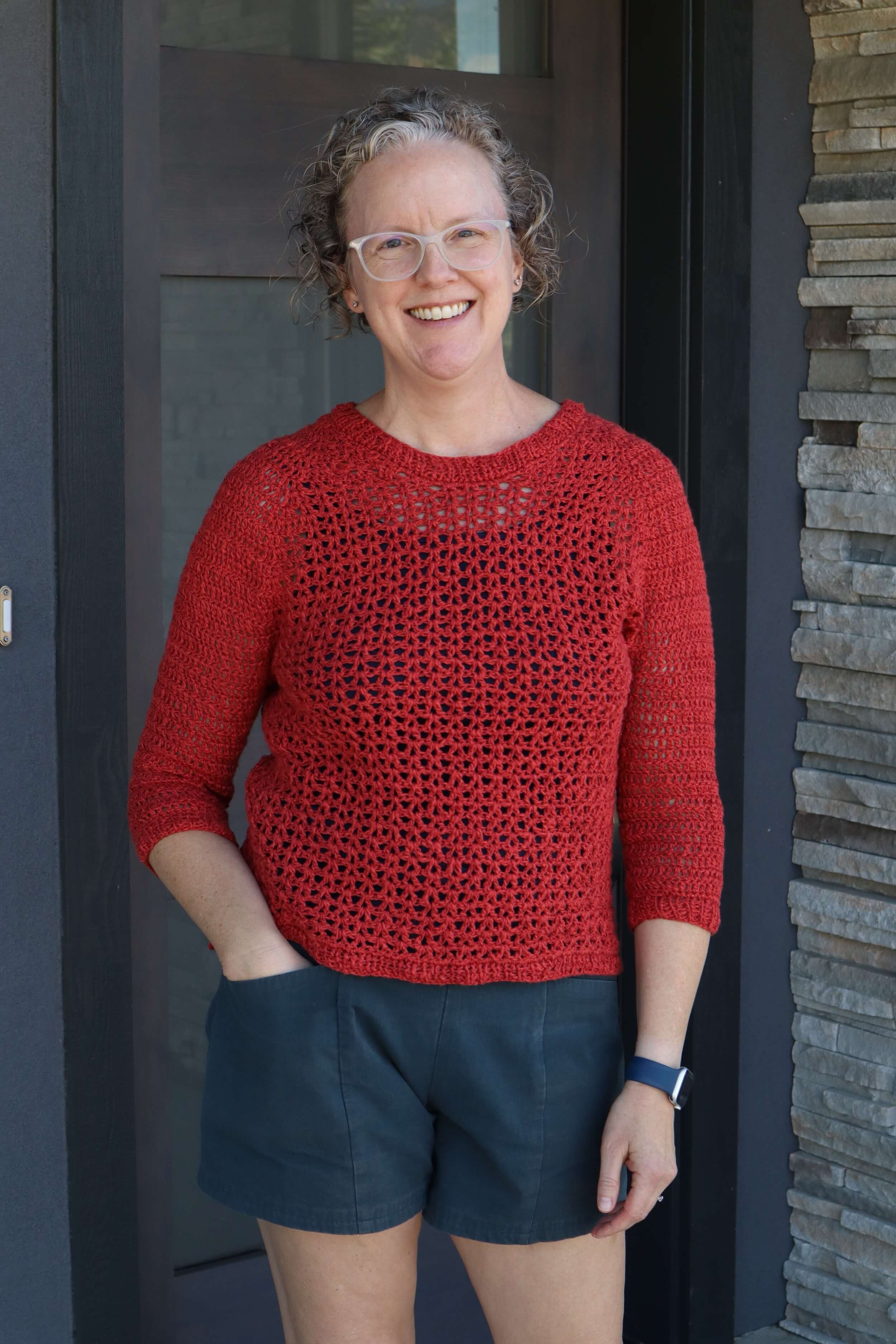 A woman stands in front of a doorway with her hand left hand in her pocket of her blue shorts. She wears a read sweater that is a mesh construction for the main body and a denser crochet stitch for the sleeves. She's got blond hair that is cut in a bob and is wearing white framed plastic glasses. She's smiling at the camera.