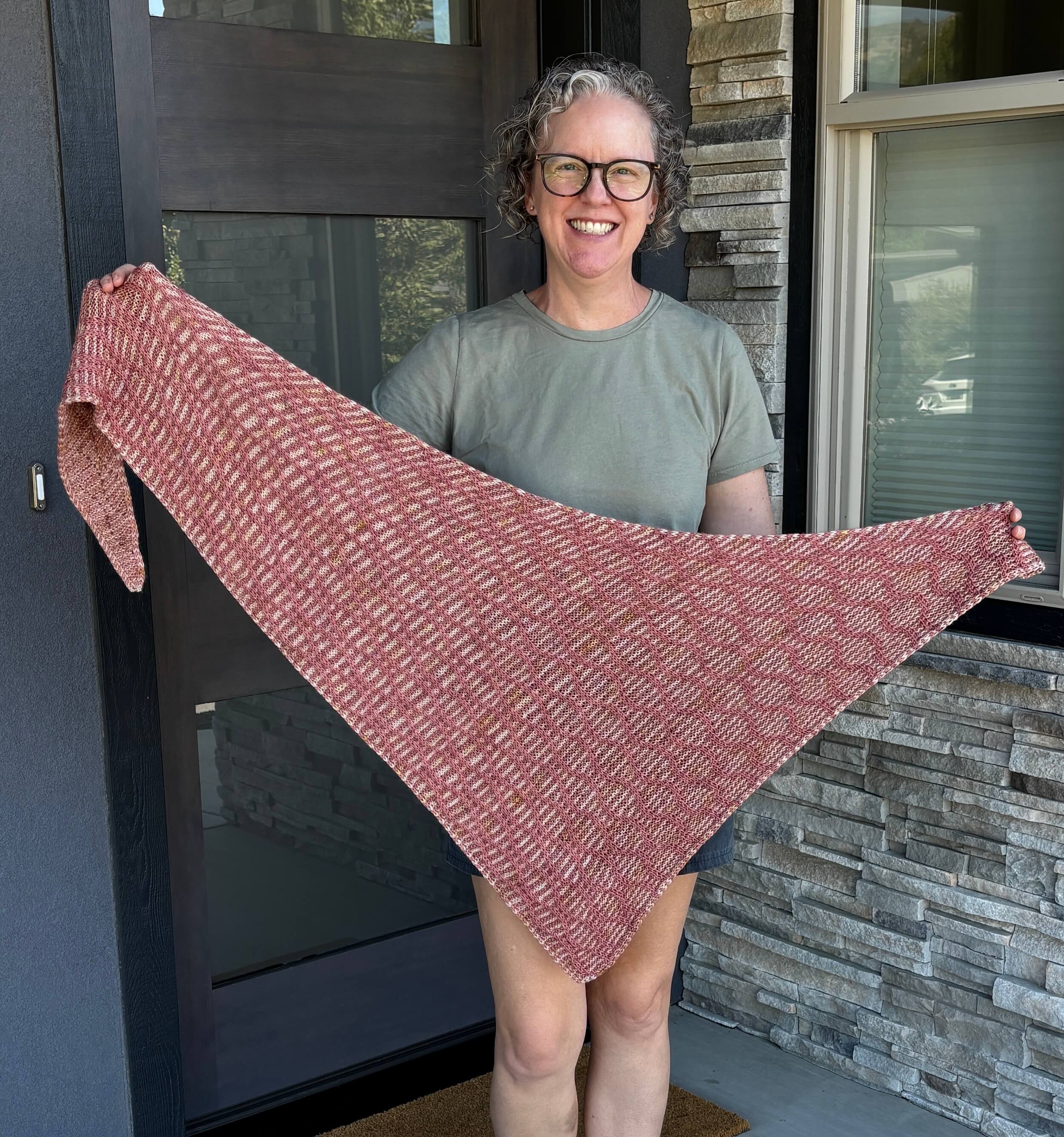A woman stands in front of a doorway wearing a light green t-shirt. She's holding a triangular shawl out in front of her that is rose colored with a vining pattern of stitches going up it. The background color against which the vines sit is a light cream color and rose stripe. She's got curly dirty blond hair and dark brown glasses on and is smiling.