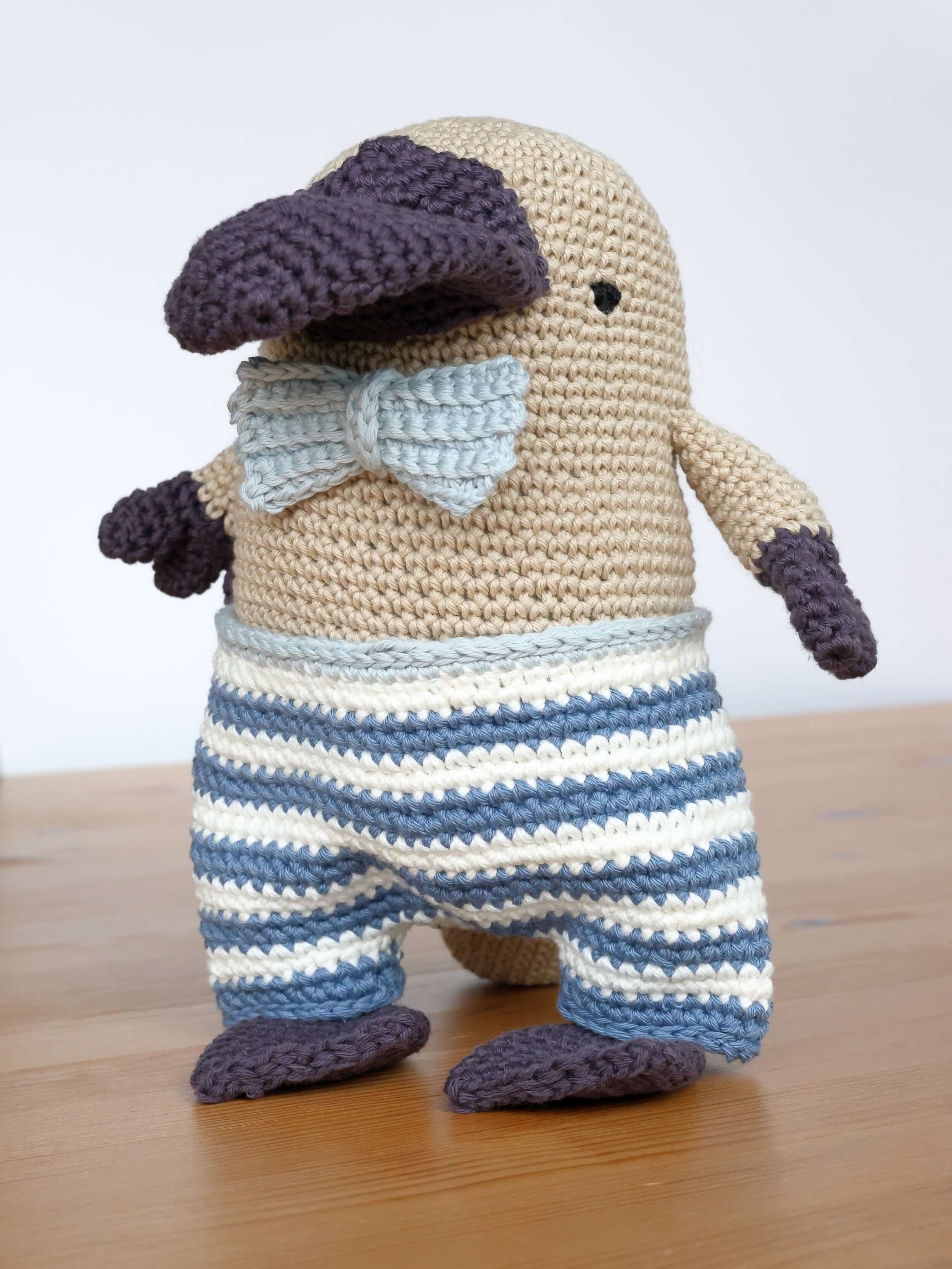 A crocheted figure stands against a white background on a brown table. He's a platypus that is standing up and he's wearing a light blue bowtie with light blue, dark blue, and white striped trousers. His body is a light cream color and his hands, feet, and beak are a very dark purple.