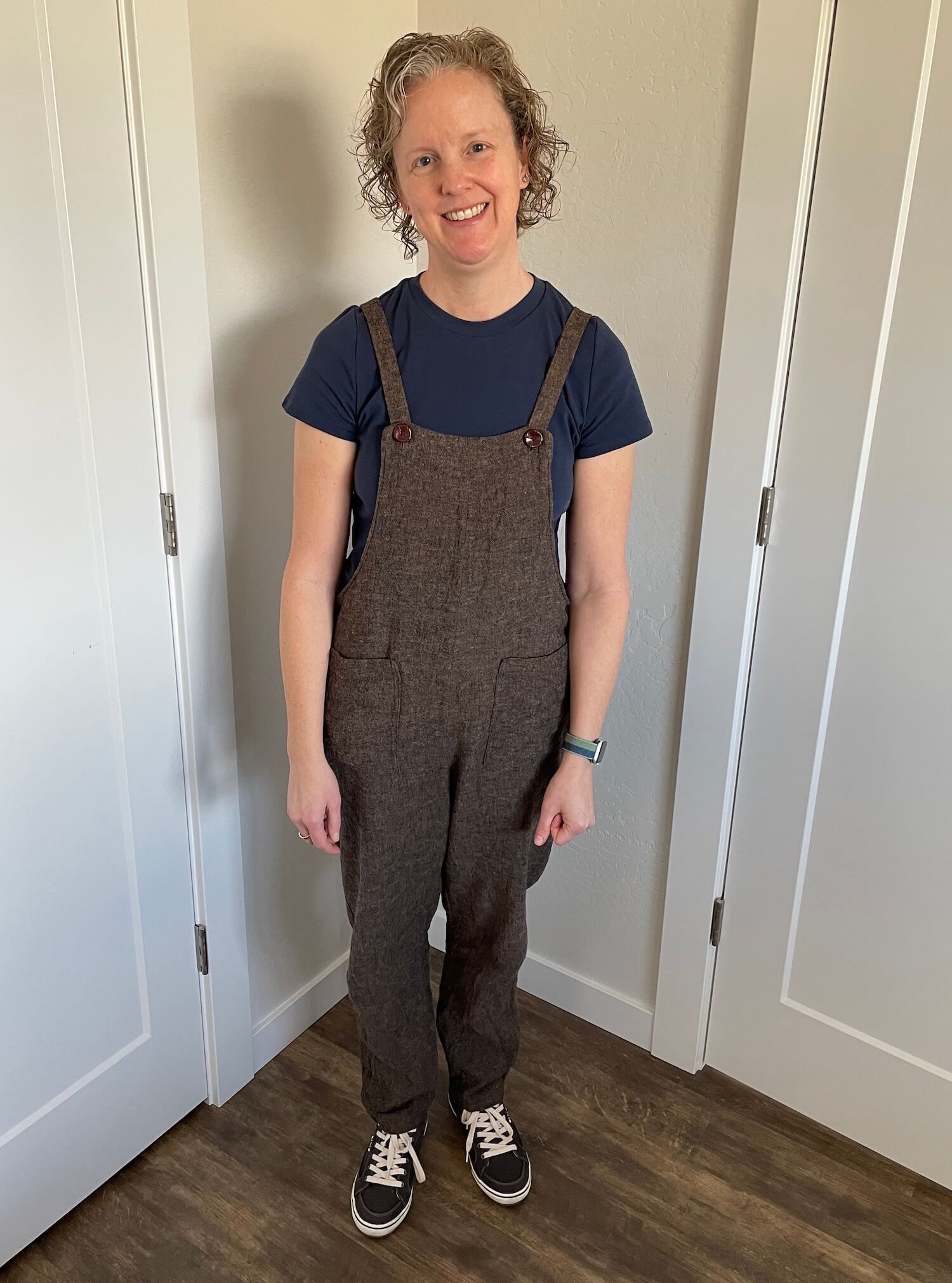Me, standing in front of a corner of a room with doors on either side, wearing brown overalls with two patch pockets and buttons for the straps, a blue t-shirt underneath, and gray tennis shoes on my feet, my head is tilted to one side.
