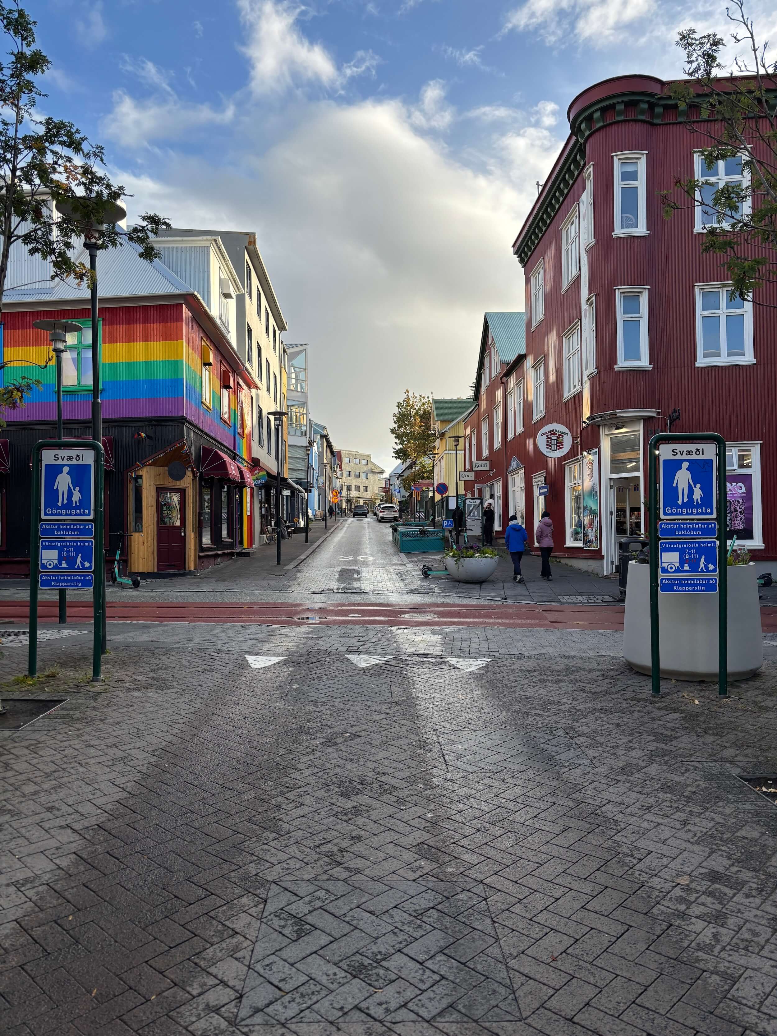 Looking up a street to an intersection that is lined by small buildings, one of which is rainbow colored on the corner, people can be seen walking on the sidewalk to the left, there are signs in Icelandic noting pedestrian walkways, and the skyy looks large with big puffy white clouds against clear blue.