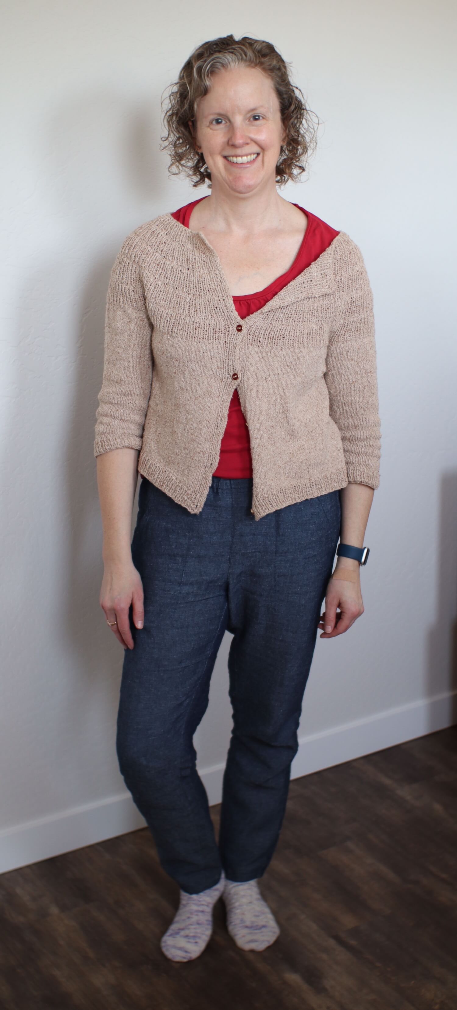 Me standing against a white wall with a cream colored cardigan with 3/4 sleeves partially buttoned, a red scoop neck t-shirt, and a pair of denim blue linen pants.