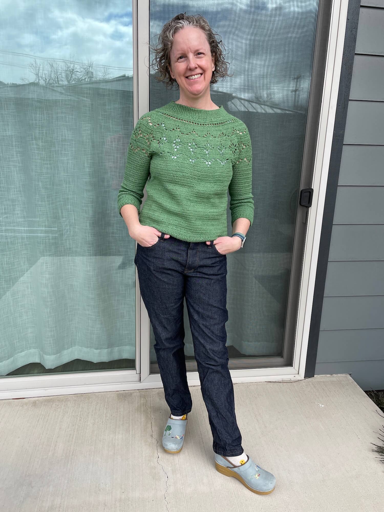 Me, in a green crochet sweater, my hands in the pockets of my jeans, standing in front of a large slider window with my left leg out and my head slightly tilted, wearing dark denim jeans.