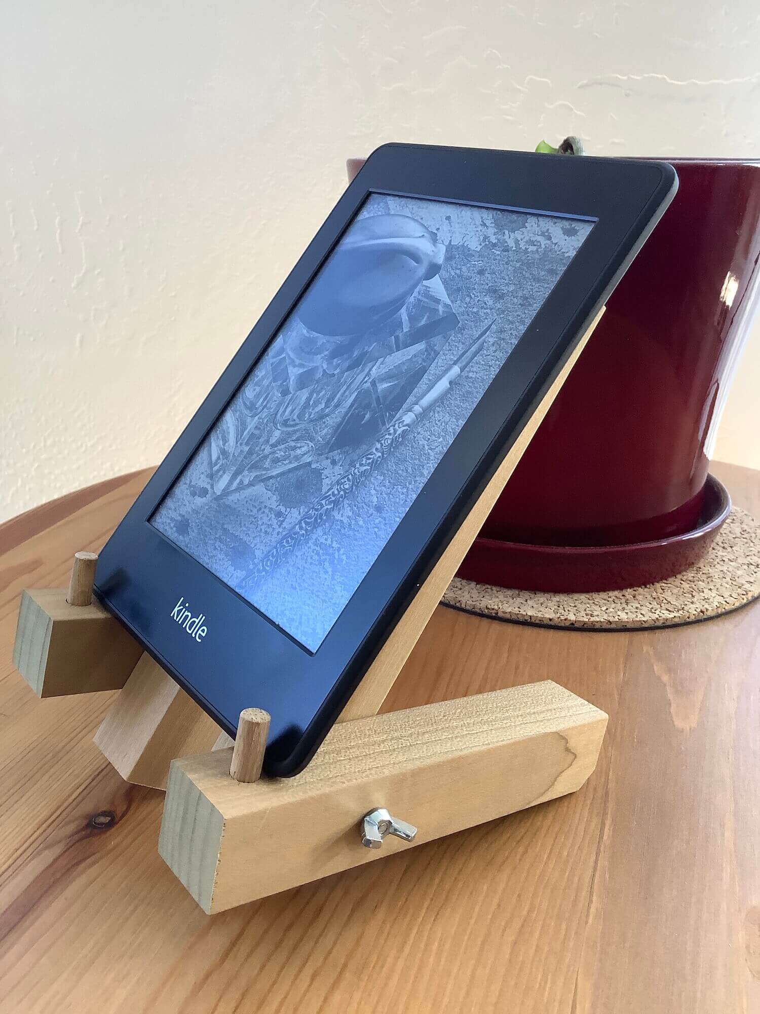 a kindle paperwhite sitting on a custom wood stand with a plant in a red pot in the background