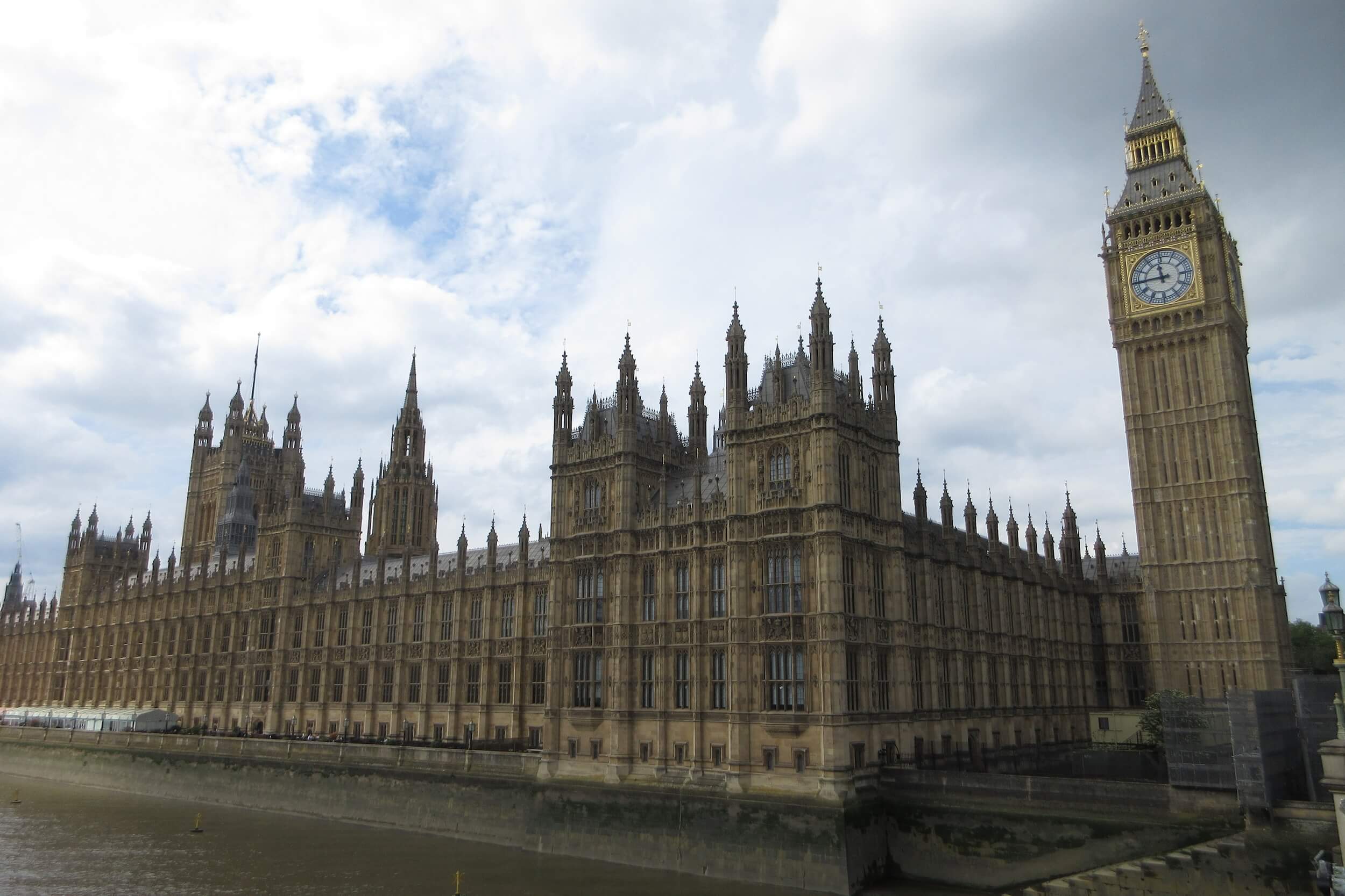 Big Ben in London is on the right with Westminster along the left and you can see the Thames in front of it. The sky is full of puffy white clouds above the buildings.