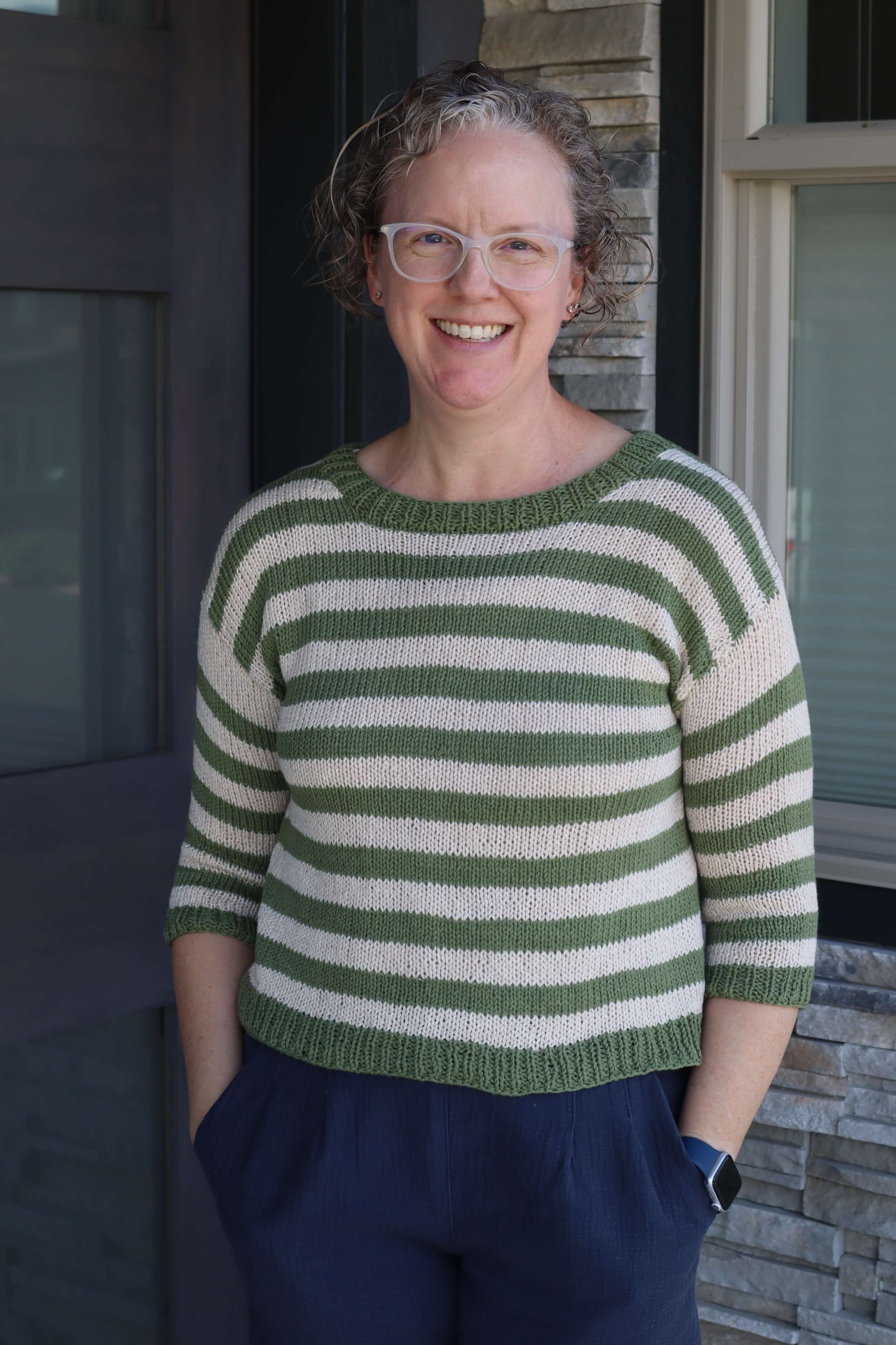 A woman stands in front of a door and window, with stone surrounding the window. She's wearing an olive green and cream striped sweater with three quarter sleeves, she's got blond curly hair, is wearing glasses that are a white plastic frame, and smiling. Her hands are in the pockets of her shorts.