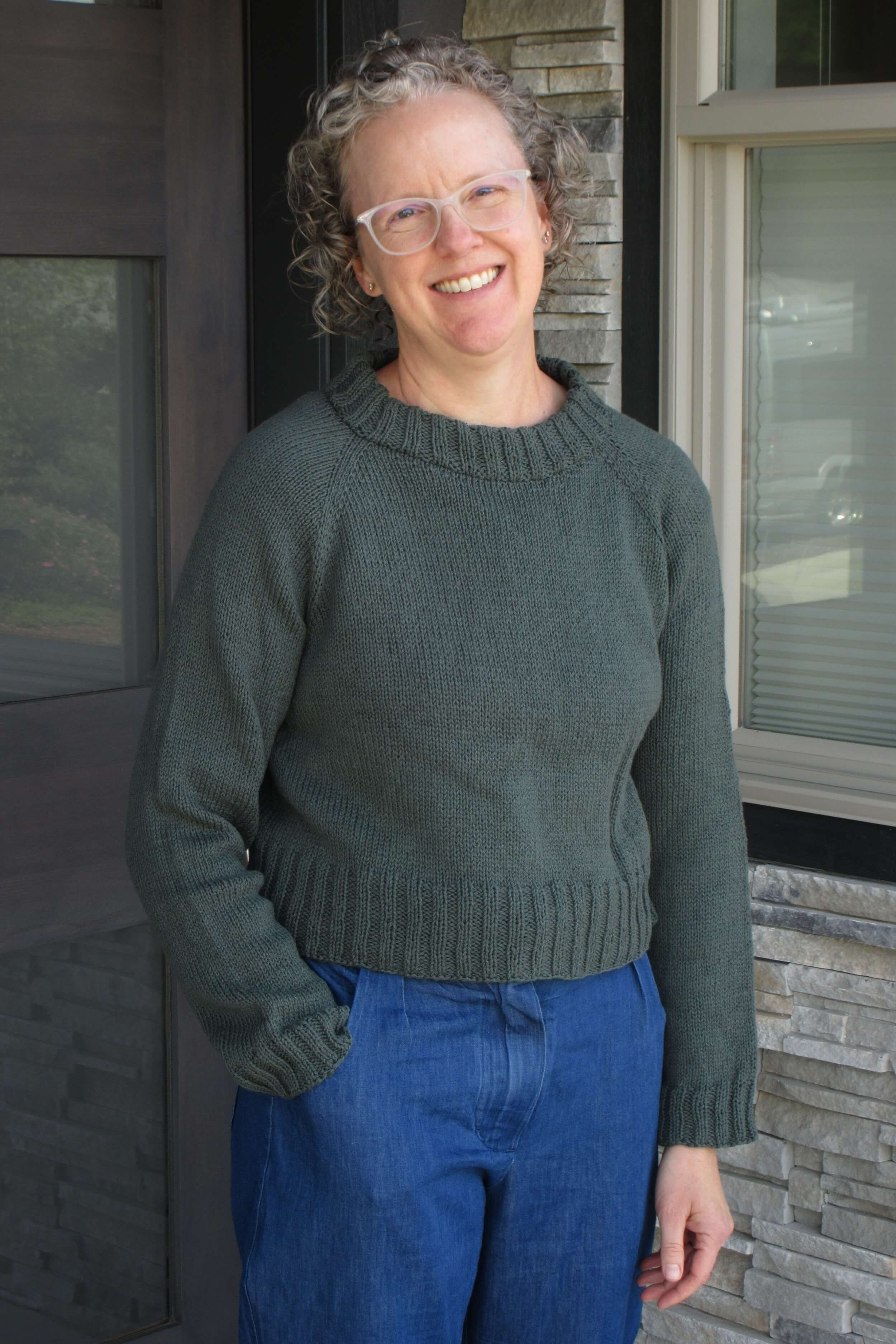 A woman stands in front of a doorway in a dark green sweater that is plain knit stitch all over. She's wearing white plastic frame glasses and has curly hair with gray at the front. She's smiling and her head is tilted to the left, one hand in the pocket of her trousers, the other straight at her side. The trousers are a denim color.