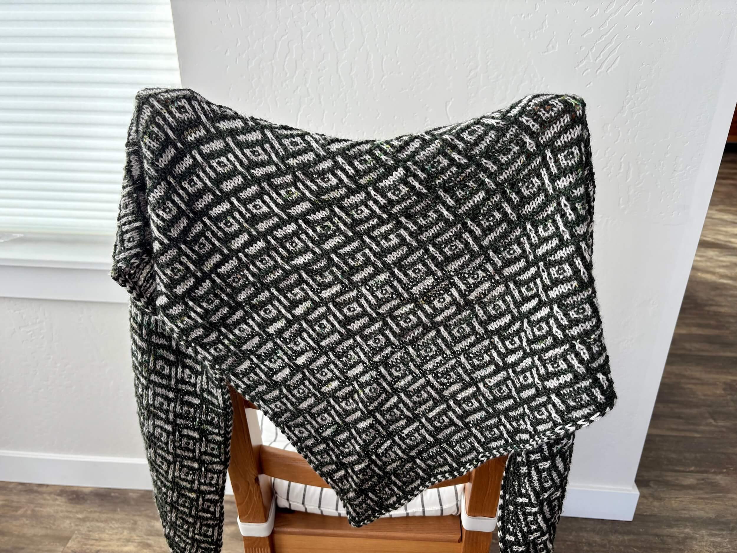 Over the back of a chair that is in front of a light gray wall, a triangular shawl hangs that is dark green and gray. The pattern on the shawl is angular lines of color that make small squares and rectangles. The chair is facing away from the viewer and there is a striped cushion on the seat. To the left of the picture you can see a shade over a window.
