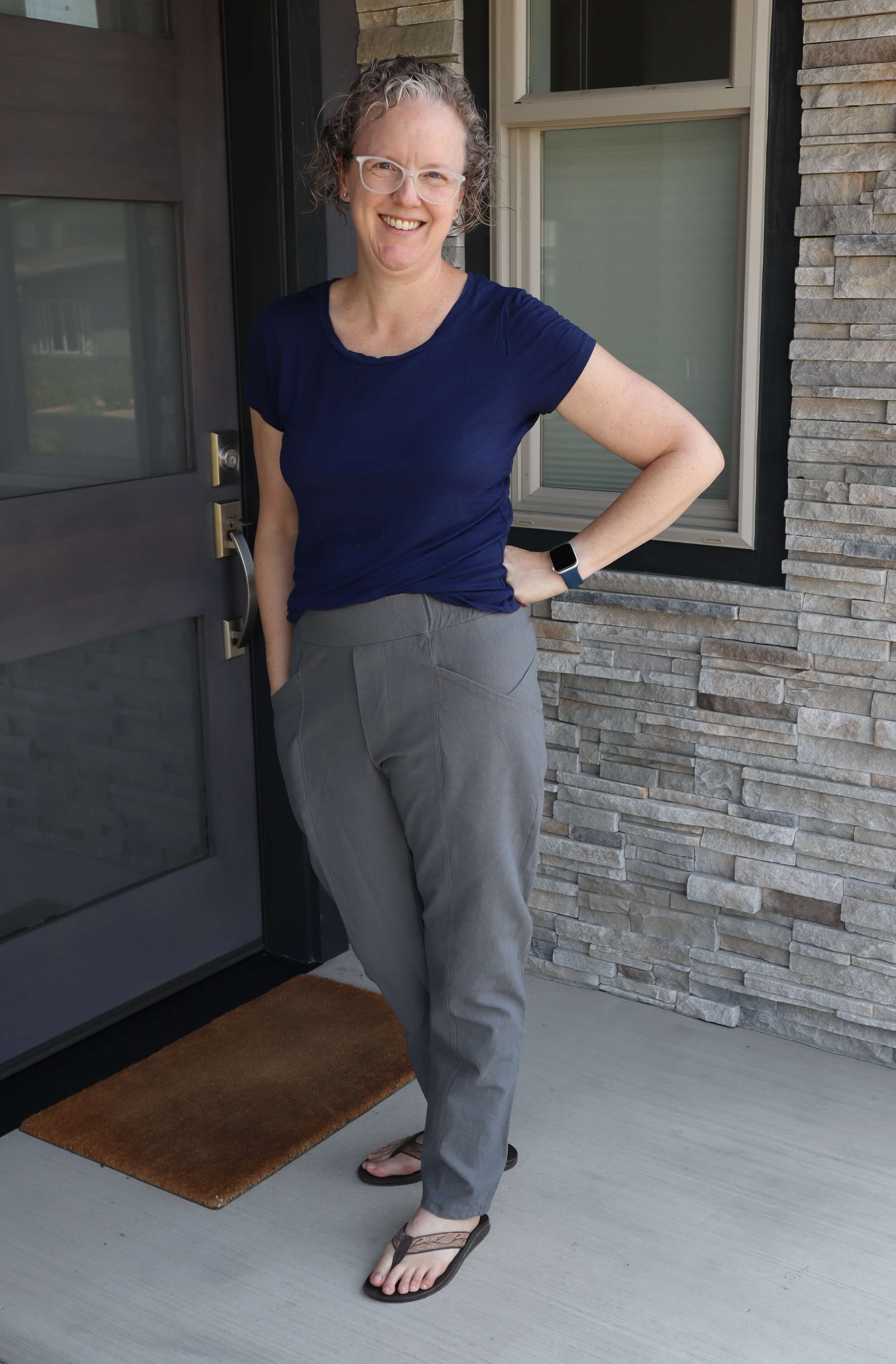 A woman stands in front of a door and window, turned slightly towards her left, she's got one hand in her left pocket and the other is resting on her hip. She's wearing a blue scoop neck t-shirt, gray high waisted pants with side pockets that slash down, and brown flip flops. She's smiling, wearing white plastic framed glasses, and has her head tipped slightly to the her right.
