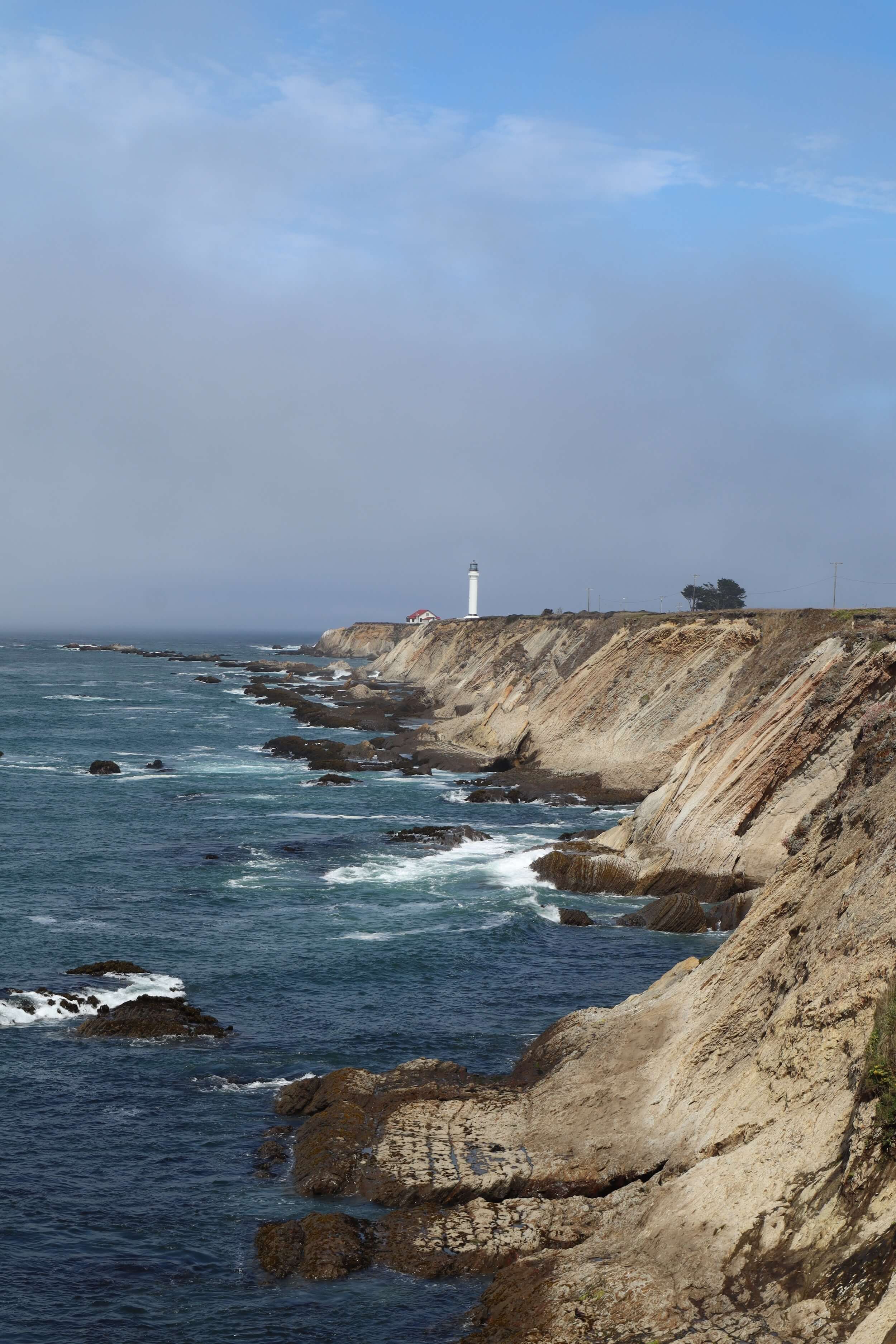 The sky is large and there is blue to the top right but also gray, foggy clouds. A rocky cliff that isn't too high is scene leading out to a point where a white lighthouse sits small in the distance, to the right of the lighthouse is a tree.