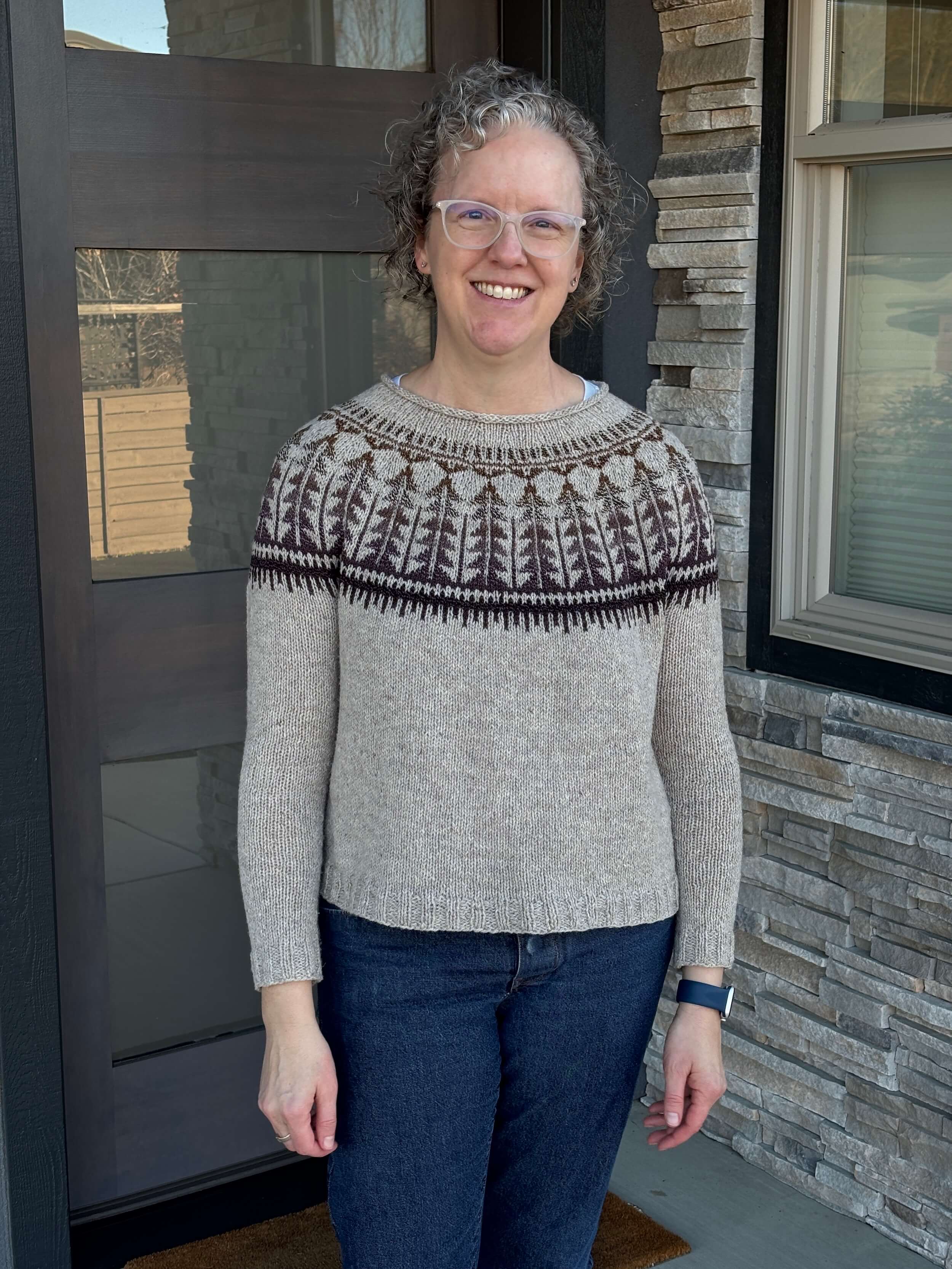 A woman stands in front of a blue door with a window to the side that's surrounded by white and gray rock. She's got curly blond/brown hair that is chin length and is wearing white plastic glasses frames. She's smiling. She's wearing an oatmeal colored sweater that has a colorwork design done in a brown that slowly changes to purple and it goes on and it's a series of triangle and lines across the yoke.