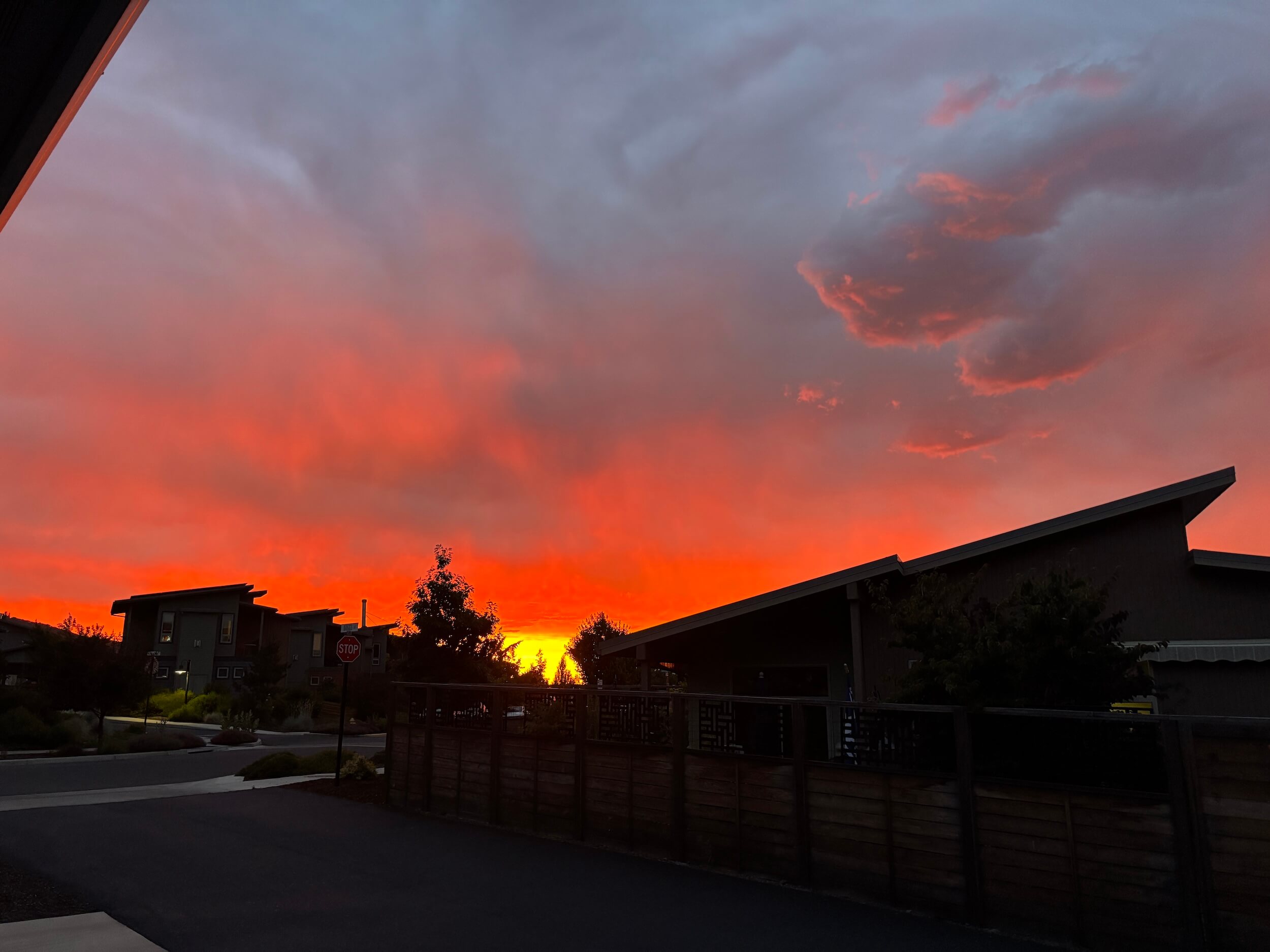 Rooftops are dark and in the foreground with the sky a fire red color and at the center of the photo, between some trees and fence lines, there is a bright gold square as the sun sets.