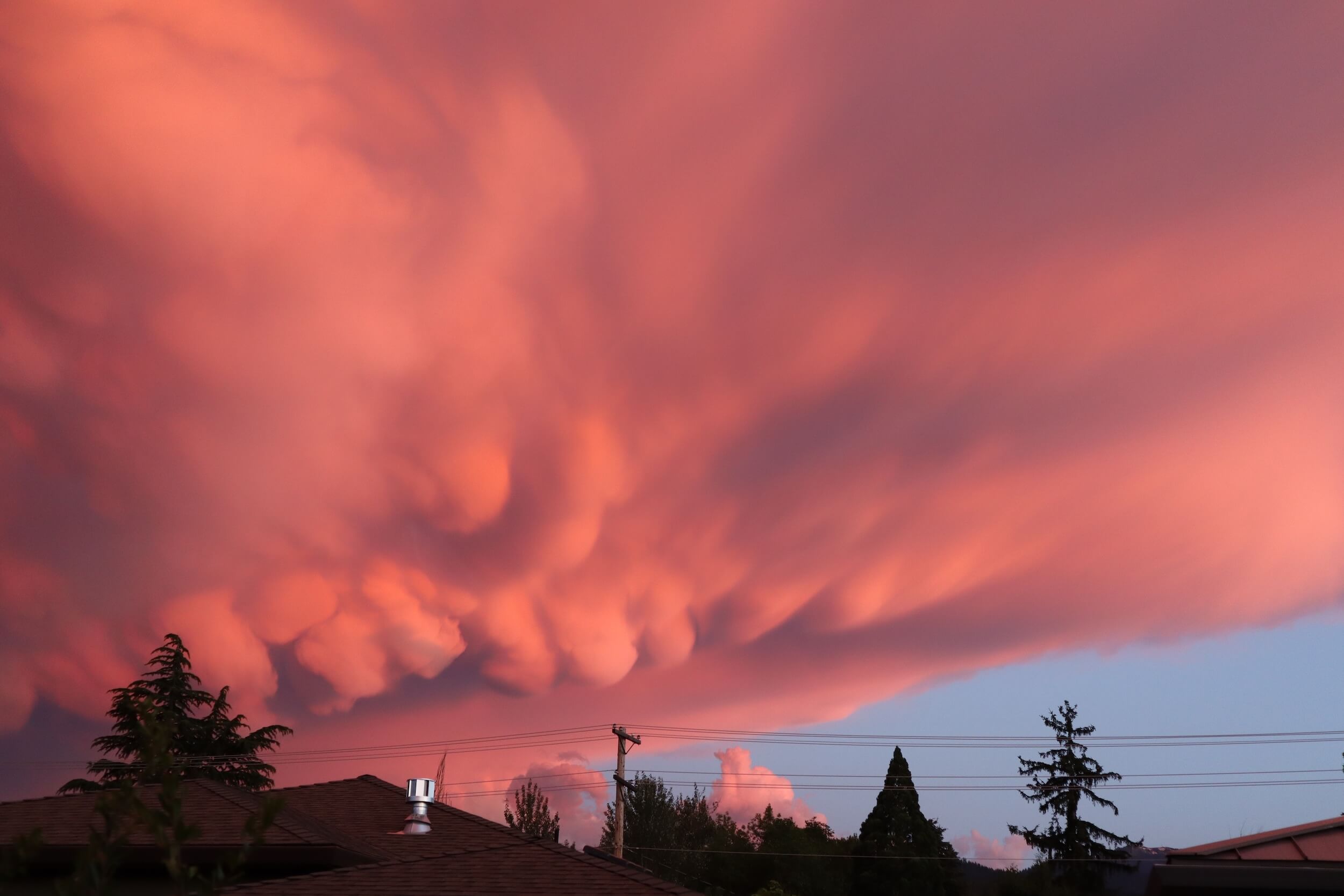 The sky is an orange pink color, with the clouds looking puffy like a marshmallow, on the bottom right there is blue sky and along the bottom edge there are some tree tops, a roof top, and electric lines coming off a ple.