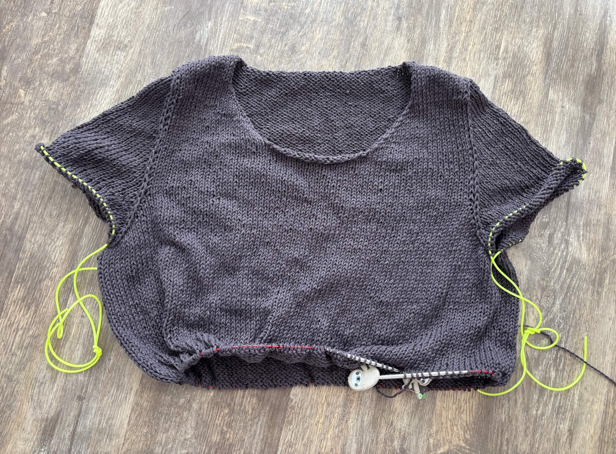 A partially knit sweater is lying flat on brown floorboards. It has bright neon green holders holding the stitches for the sleeves and the body is on the needles, with sloth needle stoppers on the ends. There is no collar yet and it's a dark, warm gray color.