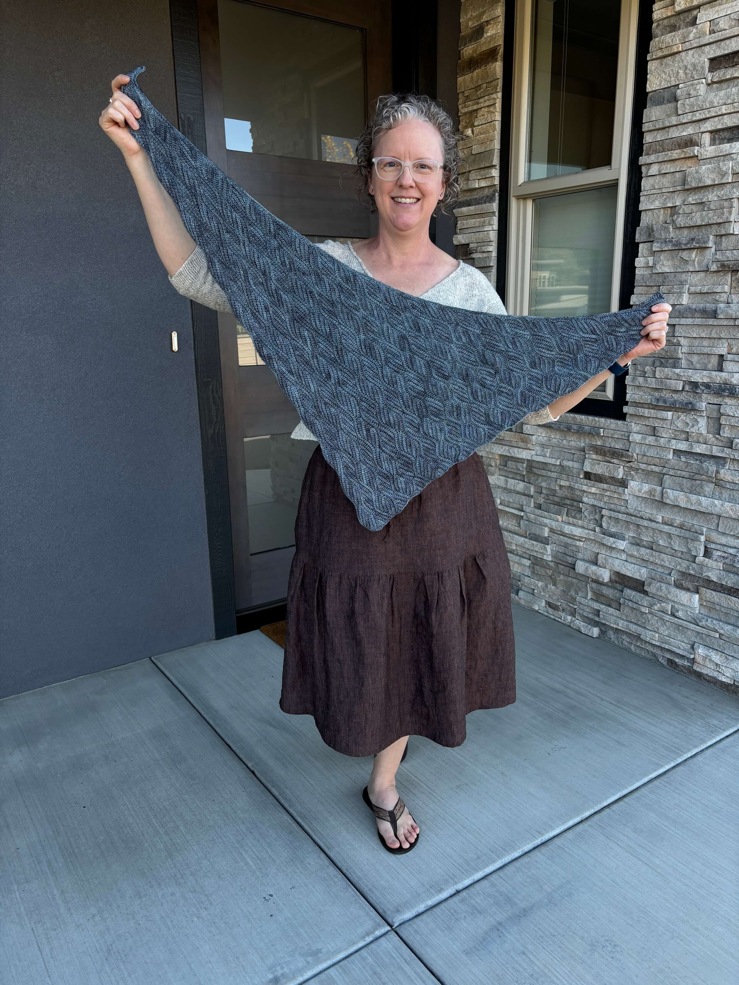 A woman stands in front of a doorway with a window to her left that's sitting in a stone wall. She's holding out a traingle shaped shawl that has the rhythm of waves in the design or small oval wedges. She's wearing a brown two tiered skirt and flip flops with a cream sweater somewhat visible behind the shawl. She's got on glasses with a white plastic frame and her hair is curly and she's smiling.