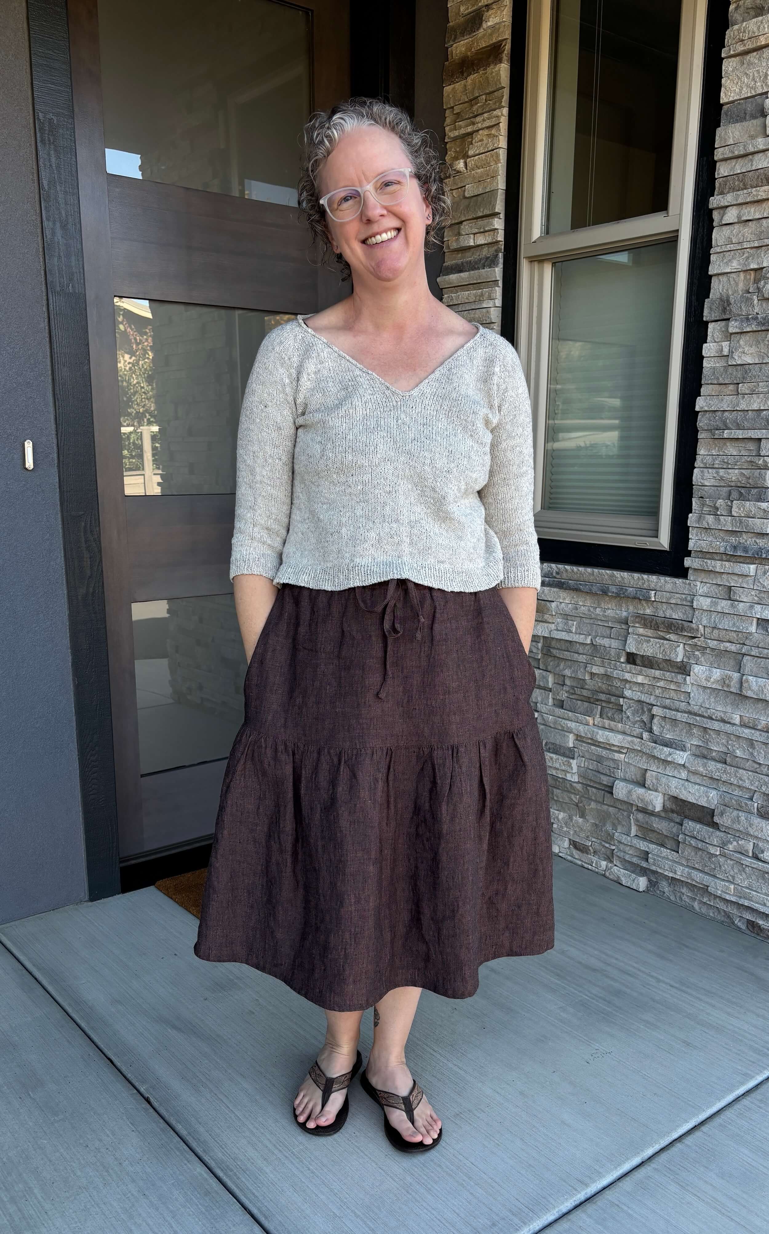 A woman stands in front of a doorway wearing a cream colored v-neck sweater and a brown tiered linen skirt. She's smiling, has curly blond/brown hair that is chin length and wears white plastic frame glasses.