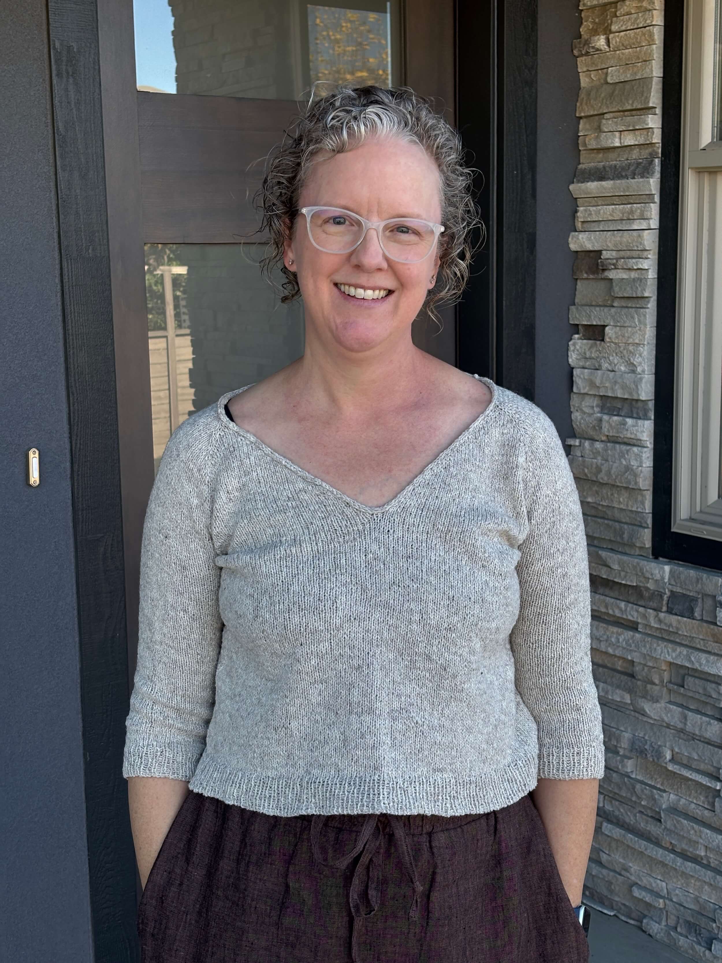 A woman stands in front of a doorway wearing a cream colored v-neck sweater and she is visible only from mid chest up. She's smiling, has curly hair about chin length and wears glasses with a white plastic frame.